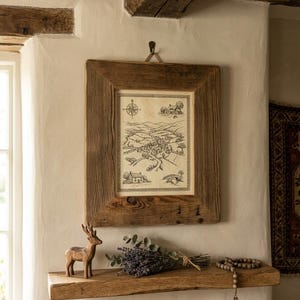 May include: A rectangular wooden frame containing a black and white line drawing of a rural landscape with a compass rose, houses, and hills. Below the frame, a wooden shelf holds a carved wooden deer, dried lavender, and wooden prayer beads.