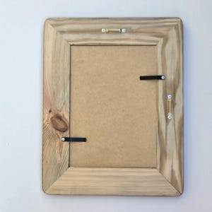 May include: A rectangular wooden picture frame shown from the back, featuring a light brown natural wood grain finish with a visible knot. The frame includes a tan backing board, two black metal swivel clips, and two gold-toned metal hanging brackets.