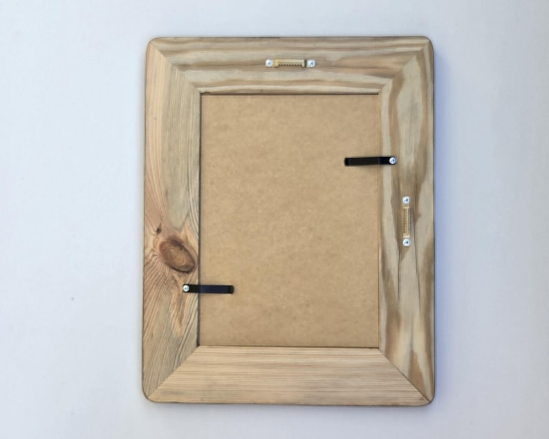 May include: A rectangular wooden picture frame shown from the back, featuring a light brown natural wood grain finish with a visible knot. The frame includes a tan backing board, two black metal swivel clips, and two gold-toned metal hanging brackets.