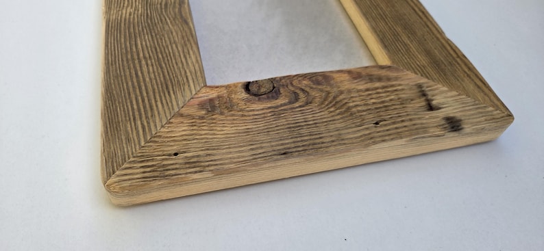 May include: A rectangular picture frame constructed from weathered, reclaimed wood with a prominent natural grain pattern. The frame features mitered corners and a visible knot in the wood on the bottom rail, showcasing a rustic, textured surface.