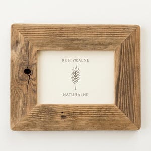 May include: A rectangular picture frame made of brown wood with a visible grain and a small knot on the left side. The frame contains a white insert featuring the text RUSTYKALNE, a line drawing of a wheat stalk, and the word NATURALNE.