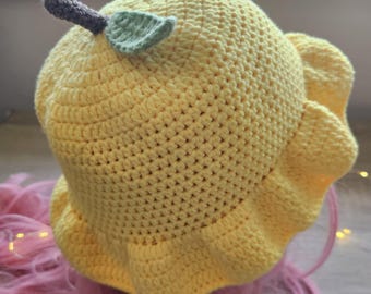Può includere: Un cappello a secchiello giallo all'uncinetto fatto a mano con un design a limone. Il cappello presenta un gambo marrone e una foglia verde. L'orlo ha un bordo a balze. Il cappello è mostrato su una parrucca rosa.