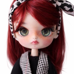 Puede incluir: Una muñeca personalizada con cabello rojo y ojos verdes. La muñeca lleva un chaleco a cuadros blanco y negro sobre una blusa negra y una diadema estampada en blanco y negro. La muñeca tiene pecas y mejillas rosadas.