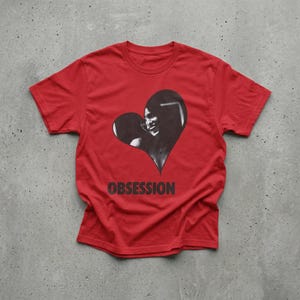 Könnte beinhalten: Ein leuchtend rotes Kurzarm-T-Shirt mit einer schwarz-weißen Grafik einer Person innerhalb einer herzförmigen Kontur. Unter der Herzgrafik ist das Wort OBSESSION in fetten, schwarzen Großbuchstaben im Brustbereich aufgedruckt.