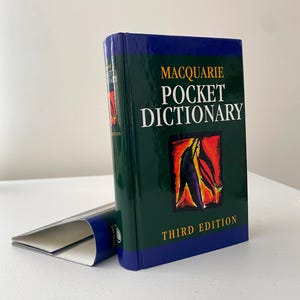 Peut inclure: Un livre relié vert et bleu intitulé "Macquarie Pocket Dictionary" avec une étiquette de troisième édition. La couverture présente une peinture abstraite d'une feuille rouge, orange et violette.