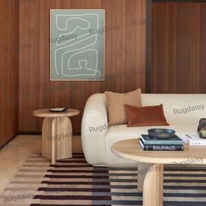 K&ouml;nnte beinhalten: Ein modernes Wohnzimmer mit cremefarbenem Sofa, braunen Kissen und einem gestreiften Teppich. Ein runder Holztisch mit B&uuml;chern und einer Schale. Ein gerahmtes abstraktes Kunstwerk h&auml;ngt an der holzget&auml;felten Wand. Zwei h&ouml;lzerne Beistelltische sind ebenfalls sichtbar.