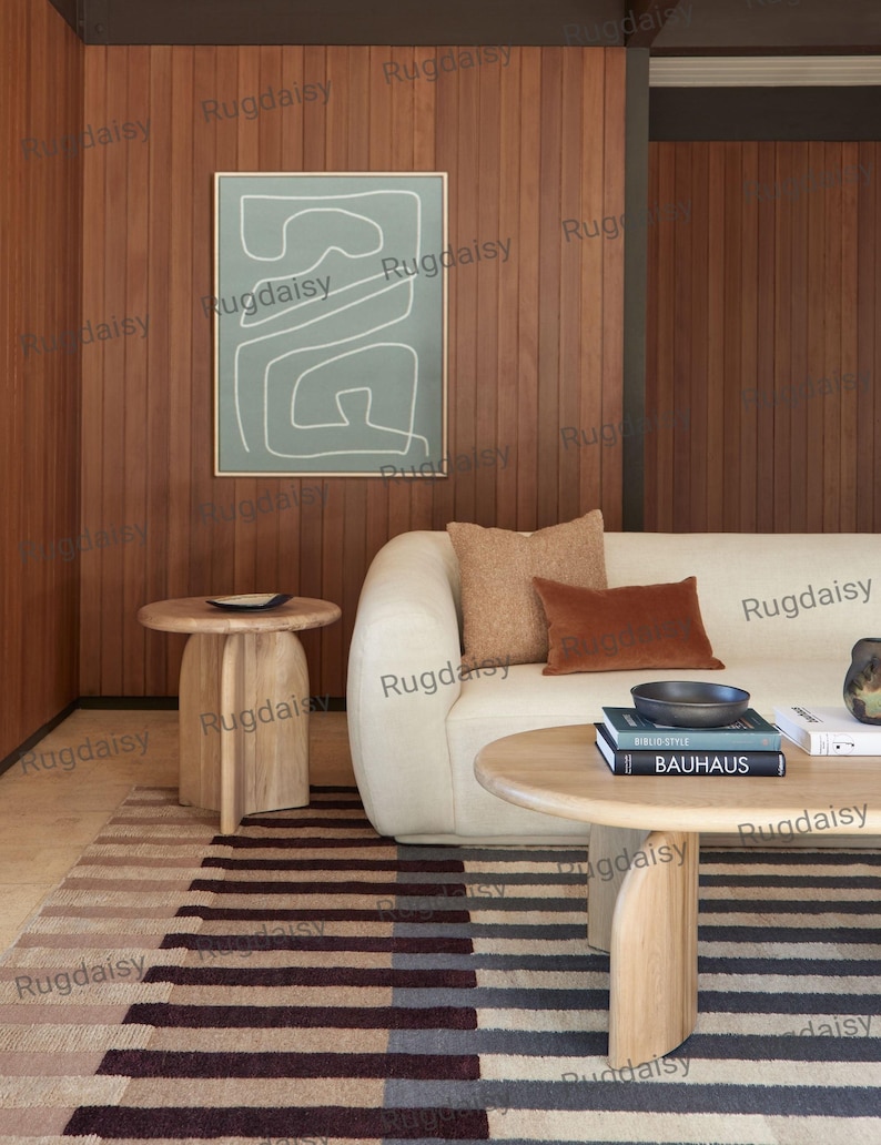 K&ouml;nnte beinhalten: Ein modernes Wohnzimmer mit cremefarbenem Sofa, braunen Kissen und einem gestreiften Teppich. Ein runder Holztisch mit B&uuml;chern und einer Schale. Ein gerahmtes abstraktes Kunstwerk h&auml;ngt an der holzget&auml;felten Wand. Zwei h&ouml;lzerne Beistelltische sind ebenfalls sichtbar.