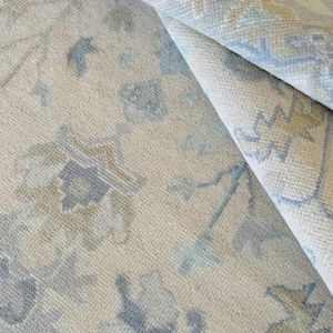May include: A rolled-up area rug with a floral pattern in shades of beige, blue, and grey. The rug's design features stylized floral motifs and geometric shapes. The rug appears to be made of a soft, woven material.