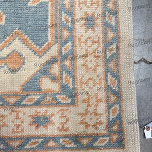 Könnte beinhalten: Nahaufnahme eines Teppichs mit geometrischem Design. Der Teppich hat einen cremefarbenen Hintergrund mit blauen und pfirsichfarbenen Mustern. Der Rand hat ein sich wiederholendes Muster aus blauen Dreiecken und pfirsichfarbenen Formen.