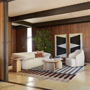 K&ouml;nnte beinhalten: Ein modernes Wohnzimmer mit einem cremefarbenen Sofa, einem passenden Sessel und einem Holztisch. Der Raum verf&uuml;gt &uuml;ber einen gestreiften Teppich in Grau-, Braun- und Beiget&ouml;nen. Zwei abstrakte schwarz-wei&szlig;e gerahmte Kunstwerke h&auml;ngen an der holzget&auml;felten Wand.