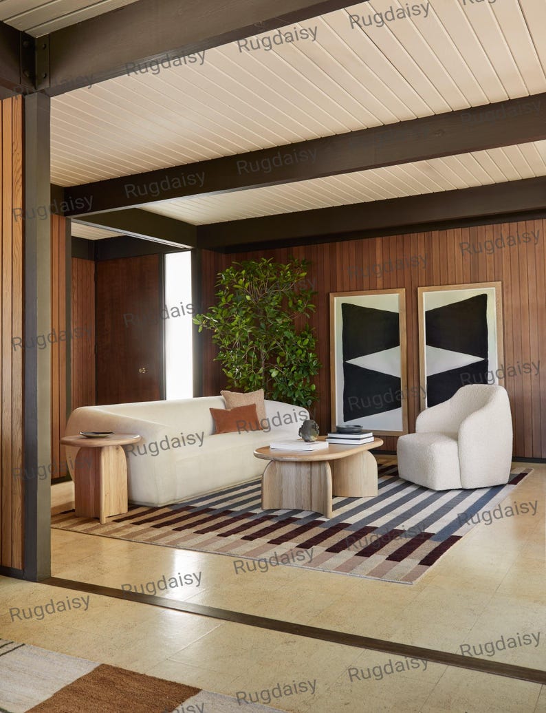 K&ouml;nnte beinhalten: Ein modernes Wohnzimmer mit einem cremefarbenen Sofa, einem passenden Sessel und einem Holztisch. Der Raum verf&uuml;gt &uuml;ber einen gestreiften Teppich in Grau-, Braun- und Beiget&ouml;nen. Zwei abstrakte schwarz-wei&szlig;e gerahmte Kunstwerke h&auml;ngen an der holzget&auml;felten Wand.