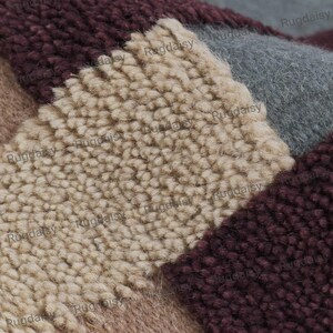 K&ouml;nnte beinhalten: Nahaufnahme eines mehrfarbigen Teppichs mit geometrischem Muster. Der Teppich hat Abschnitte in Beige, Burgunderrot, Grau und Braun. Die Textur wirkt weich und pl&uuml;schig, mit einem hohen Flor. Der Markenname "Rugdaisy" ist im Bild sichtbar.