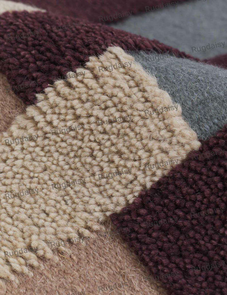K&ouml;nnte beinhalten: Nahaufnahme eines mehrfarbigen Teppichs mit geometrischem Muster. Der Teppich hat Abschnitte in Beige, Burgunderrot, Grau und Braun. Die Textur wirkt weich und pl&uuml;schig, mit einem hohen Flor. Der Markenname "Rugdaisy" ist im Bild sichtbar.