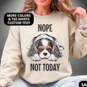 May include: Sand-colored sweatshirt with the words "NOPE" and "NOT TODAY" printed in black. A Cavalier King Charles Spaniel dog illustration is centered on the front. The sweatshirt has long sleeves and a crew neck.
