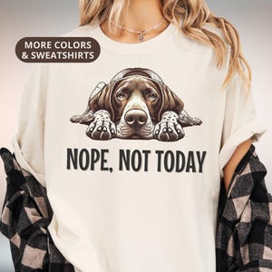 Pode incluir: Camiseta cor marfim com uma ilustração de um cão e o texto "NOPE, NOT TODAY". O cão é castanho e branco, deitado. A camisa também tem o texto "MORE COLORS & SWEATSHIRTS" e "IVORY".
