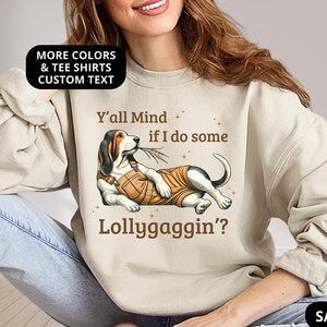 Könnte beinhalten: Sandfarbener Pullover mit einem Cartoon-Basset-Hound mit Latzhose und dem Text "Y'all Mind if I do some Lollygaggin'?" Der Pullover hat auch ein schwarzes Oval mit der Aufschrift "MORE COLORS & TEE SHIRTS CUSTOM TEXT".