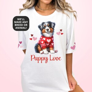 May include: White t-shirt featuring a watercolor illustration of a puppy wearing a red sweater with white hearts. The text "Puppy Love" is below the puppy. The shirt also has the text "WE'LL MAKE ANY BREED OR ANIMAL!"