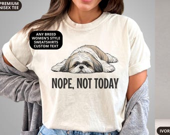 May include: An ivory short-sleeve t-shirt featuring a graphic of a brown and white Shih Tzu dog lying down with its head resting on its paws. Below the dog, the text reads "NOPE, NOT TODAY" in bold black capital letters. The shirt has a crew neckline.