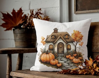 Puede incluir: Cojín decorativo blanco con una ilustración en acuarela de una cabaña con tejado de paja, rodeada de calabazas y follaje otoñal. El cojín está colocado sobre un banco de madera, evocando un tema otoñal acogedor.