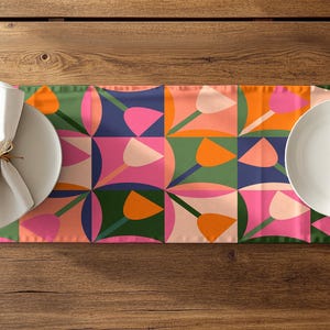 Könnte beinhalten: Ein farbenfroher Tischläufer mit einem geometrischen Blumenmuster in Pink, Orange, Grün und Blau. Zwei weiße Teller mit gefalteten Servietten und goldfarbenem Besteck sind auf dem Läufer platziert, der auf einem Holztisch liegt.