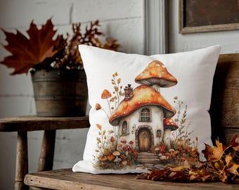 Puede incluir: Cojín decorativo blanco con una ilustración caprichosa de una casa de setas con tapas naranjas y marrones. El cojín está colocado en un banco de madera con hojas de otoño y un cubo de metal con follaje otoñal en el fondo.