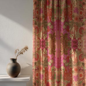 Op de afbeelding: Een decoratief gordijn met een herhalend bloemenpatroon in roze, oranje en goud. Het ontwerp omvat bloemen, palmbomen en geometrische vormen. Het gordijn hangt voor een witte muur, met een vaas en gedroogde bloemen.