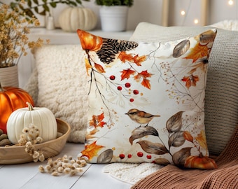 May include: A cushion with a fall theme. The cushion features a watercolour design of autumn leaves, berries, and a small bird. The colour palette includes shades of orange, brown, and cream. A pumpkin and other fall decorations are in the background.