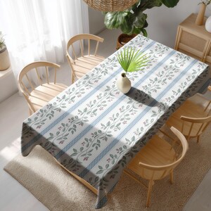 May include: A rectangular tablecloth with a floral pattern and blue stripes covers a wooden table. The floral design features green leaves and small red berries. The table is surrounded by wooden chairs.