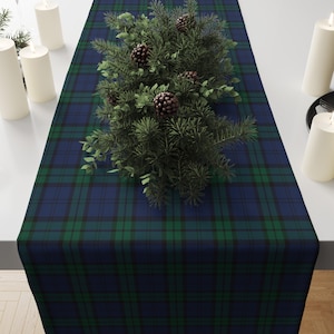 May include: A navy blue and green plaid table runner with a central arrangement of pine branches and pinecones. The runner is rectangular and placed on a white surface, with white candles on either side.