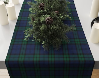 May include: A navy blue and green plaid table runner with a central arrangement of pine branches and pinecones. The runner is rectangular and placed on a white surface, with white candles on either side.