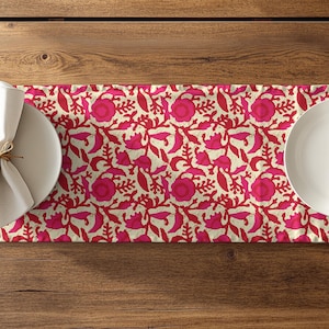 May include: A floral table runner with a cream background and a pattern of pink and red flowers and leaves. Two white plates with folded napkins and silverware are set on the runner, ready for a meal.