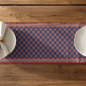 May include: A table setting featuring a navy blue and red patterned table runner with two white plates, napkins, and cutlery. The runner has a repeating floral design and is set on a wooden table.
