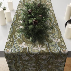 May include: A decorative table runner with a green and white floral pattern. The runner is adorned with a centerpiece of pine branches and pinecones. White pillar candles are placed on either side of the runner, creating a festive display.