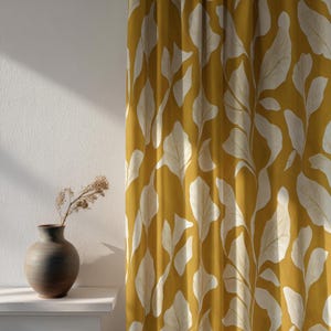 May include: Mustard yellow curtain featuring a large white leaf design. A dark grey ceramic vase with dried flowers is on a white shelf. The curtain is made of a thick fabric. The vase is approximately 20 cm tall.