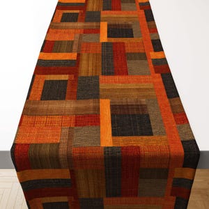 May include: A rectangular table runner with a patchwork design. The runner features a mix of rectangular blocks in shades of orange, brown, red, and black. The fabric has a textured appearance, creating a warm and inviting aesthetic.