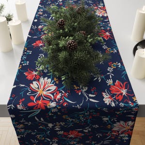 May include: A navy blue floral table runner with a vibrant pattern of red, white, and yellow flowers. The runner is decorated with a green pine arrangement and pinecones. White candles are placed on either side of the runner.