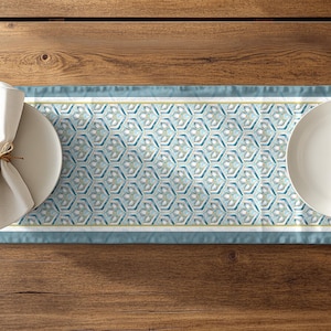 May include: A rectangular table runner with a geometric pattern in shades of blue, teal, and white. The runner is placed on a wooden table, with white plates, napkins, and silverware on either side.