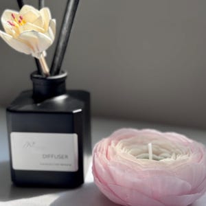 Może przedstawiać: Czarny, kwadratowy dyfuzor z trzciną i białym kwiatem, obok różowo-białej świecy w kształcie kwiatu. Świeca ma knot i stoi na białej powierzchni. Dyfuzor ma etykietę z napisem "DIFFUSER".