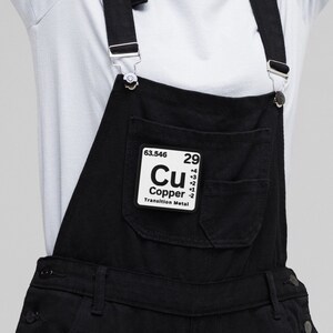 May include: A woman wearing a light blue t-shirt and black overalls with a copper element patch. The patch has the chemical symbol Cu and the atomic number 29.