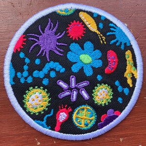 May include: A round black patch with a white border, featuring a colourful embroidered design of various microbes and bacteria. The patch is perfect for adding a quirky touch to clothing, bags, or other accessories.