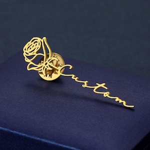 May include: Gold-toned rose pin with a delicate, cursive 'custom' inscription. The rose is detailed and the pin is attached to a backing.