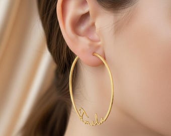 May include: Gold-colored hoop earrings with the name "Amila" in a cursive font. The earrings are large and circular, with a smooth, polished finish. The design is elegant and personalized, suitable for everyday wear or special occasions.