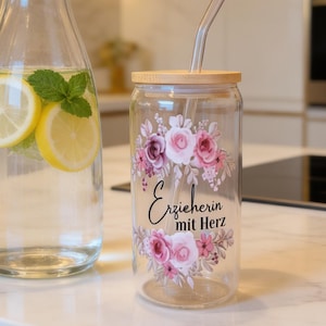 Op de afbeelding: Een heldere glazen karaf gevuld met water, schijfjes citroen en muntblaadjes, naast een glazen beker met een bamboe deksel, een glazen rietje en een bloemmotief met de zwarte tekst "Erzieherin mit Herz" op de voorzijde gedrukt.