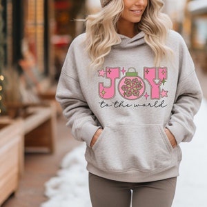 Könnte beinhalten: Ein hellgrauer Kapuzenpullover mit einer Vordertasche. Auf der Vorderseite stehen die Worte "JOY to the world" in Rosa und Grün, mit einer Weihnachtskugel-Grafik.