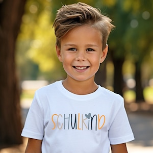 Könnte beinhalten: Ein weißes T-Shirt mit dem Aufdruck "SCHULKIND" in Orange, Blau und Grau. Das T-Shirt wird von einem lächelnden Kind getragen. Der Hintergrund ist eine verschwommene Außenszene mit Bäumen.