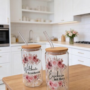 Op de afbeelding: Twee doorzichtige glazen bekers met bamboe deksels en glazen rietjes. Elke beker heeft een bloemmotief in roze en bruine tinten, met zwarte tekst "Weltbeste Erzieherin" op de ene en "Erzieherin mit Herz" op de andere beker.