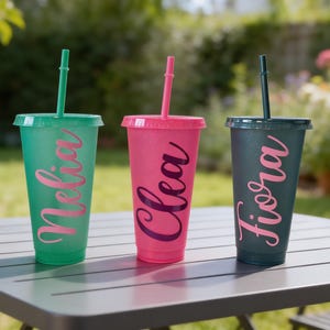 Puede incluir: Tres vasos de pl&aacute;stico reutilizables con tapas y pajitas en verde, rosa y gris oscuro. Cada vaso tiene un acabado brillante y un nombre personalizado en letra cursiva: Nelia en el vaso verde, Clea en el rosa y Fiora en el vaso de color gris oscuro.