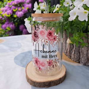 May include: A clear glass tumbler with a bamboo lid and a bent glass straw. The glass features a floral design in shades of pink and burgundy, with the black text "Hebamme mit Herz" printed in the center between the floral arrangements.