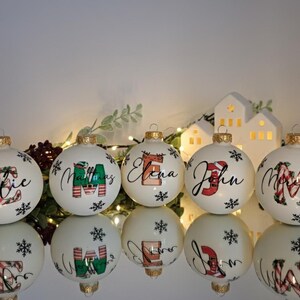 以下が含まれることがあります： 金色のキャップが付いた白いクリスマスオーナメント。それぞれに、エレガントな筆記体とお祝いのデザインで名前がパーソナライズされています。名前はさまざまな色で、雪の結晶のアクセントが付いています。装飾的な休日のアイテム。