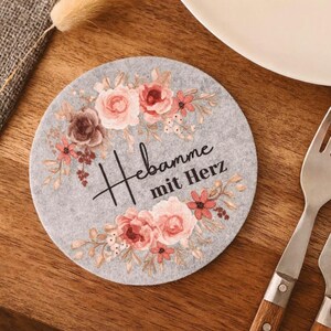 May include: A circular light gray felt coaster featuring a floral wreath design in shades of pink, peach, and brown. The text "Hebamme mit Herz" is printed in black script and serif font in the center of the floral arrangement on the coaster.